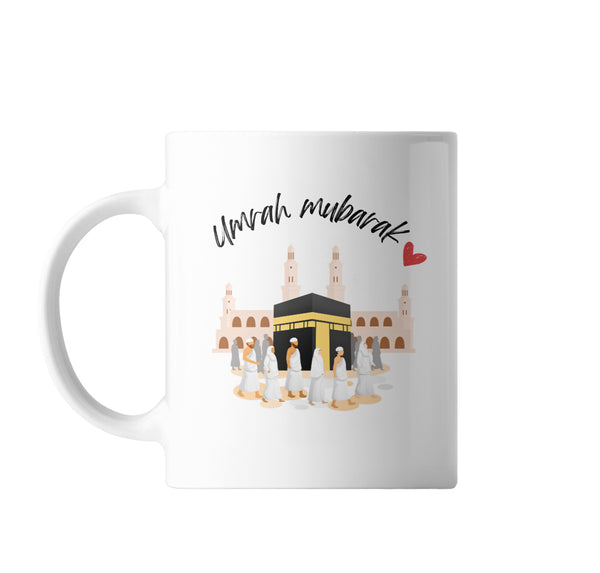 A white ceramic coffee mug featuring an illustration of the Kaaba surrounded by pilgrims and the words "Umrah Mubarak" written above in elegant script with a small red heart to the right.