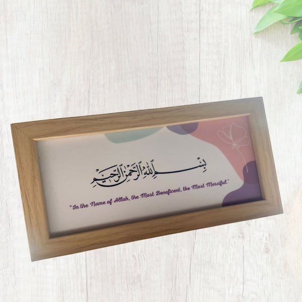 Wooden-framed desk art featuring the Bismillah in Arabic calligraphy with an English translation below, set against a soft pastel background with abstract shapes and a butterfly design. The frame is placed on a light wooden surface with a green plant partially visible in the corner.